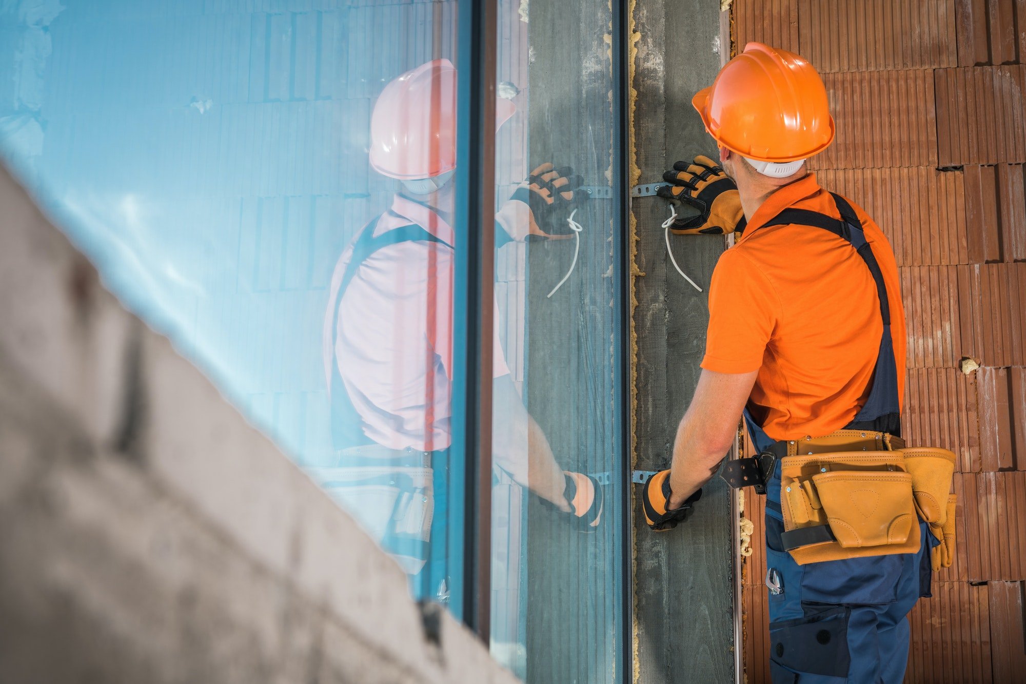 Worker in an orange hard hat and tool belt fitting a large glass window beside a brick wall