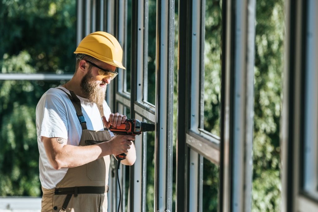Bearded builder in a yellow hard hat and safety glasses drilling a window frame