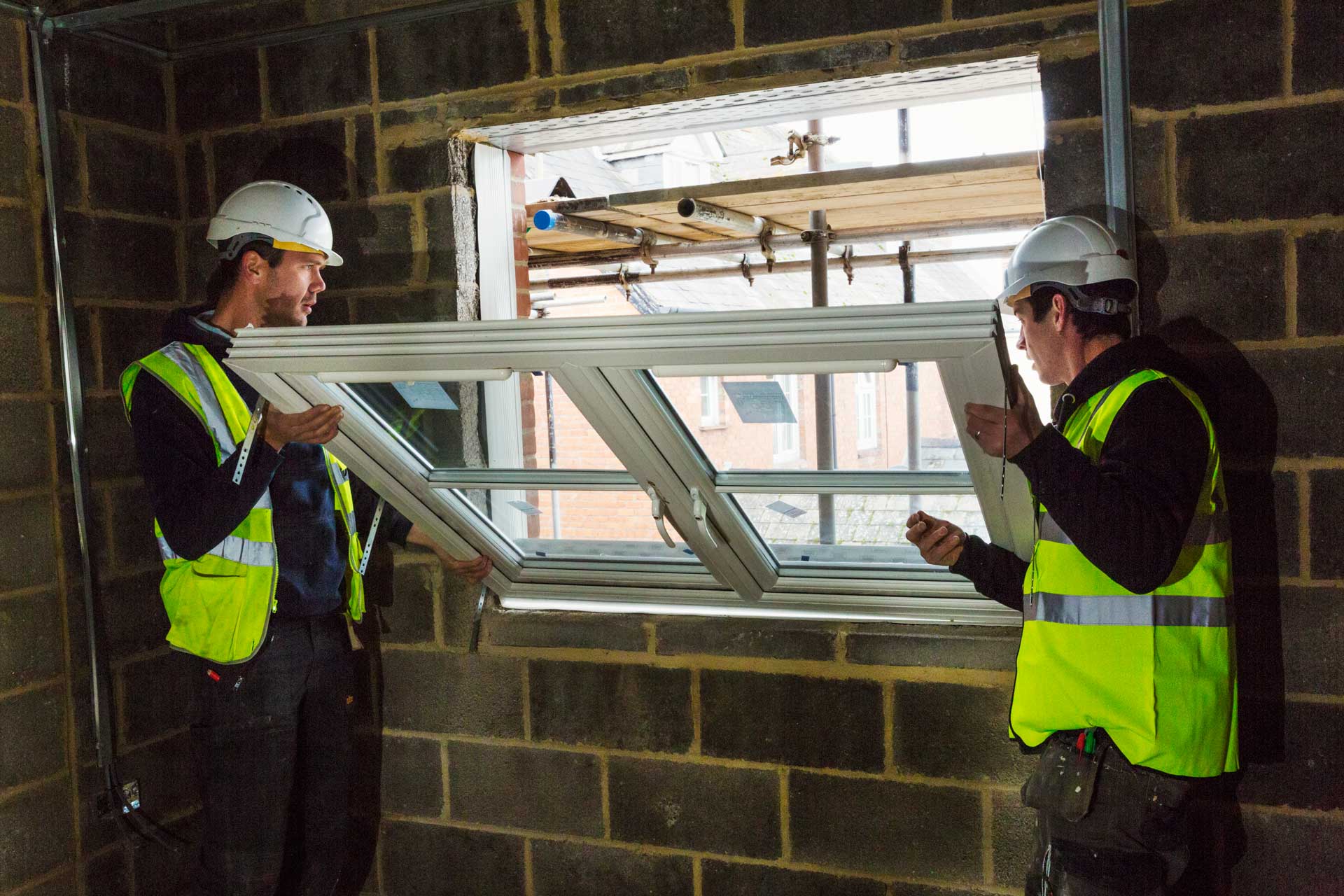 Two builders in hard hats and hi-vis vests holding a white window frame in a block wall opening